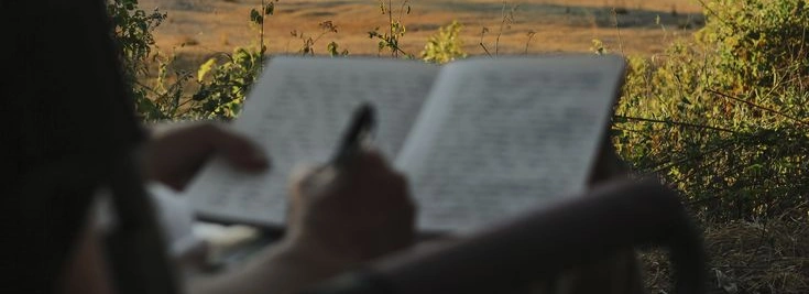 Una persona escribe en un cuaderno al aire libre frente a un paisaje de campo.