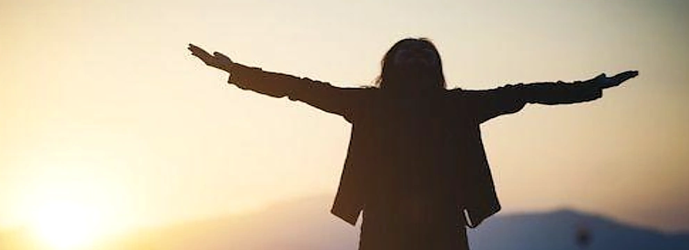 Una persona con los brazos abiertos frente a un paisaje de montañas al atardecer.